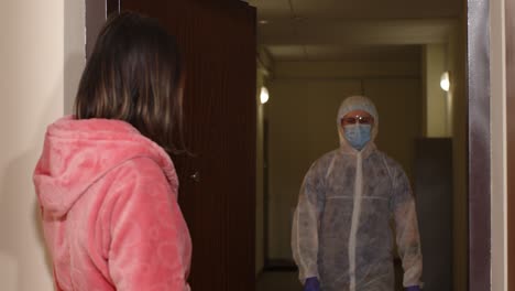 a healthcare worker in full ppe, wearing a mask and gloves, is holding a syringe and a sign that says 'covid-19 vaccination station'. a woman in a pink robe, also wearing a mask, claps at him in appreciation.