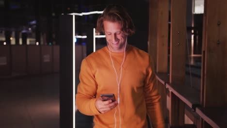 caucasian male wearing earphones and skateboard, smiling, walking, using his phone in the evening