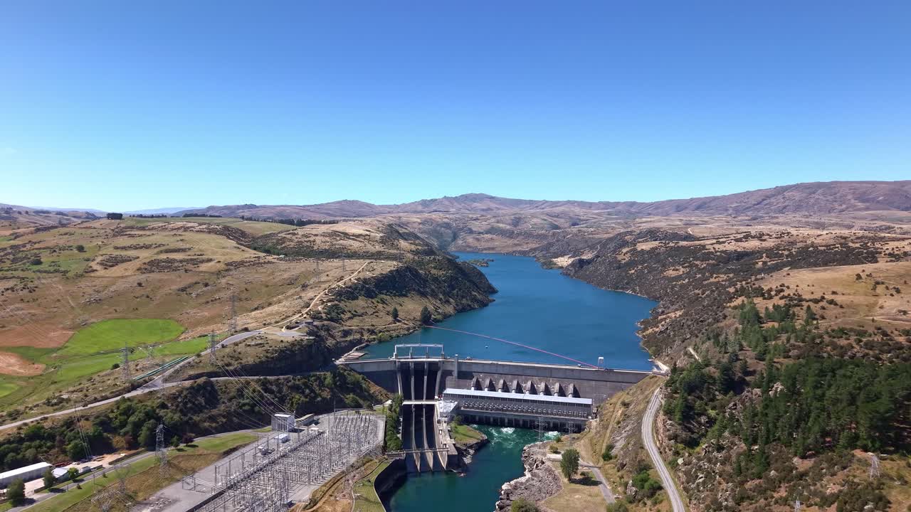 Drone flies toward Roxburgh hydroelectric dam, capturing the power station, full dam structure, and scenic Roxburgh Lake in Central Otago, New Zealand