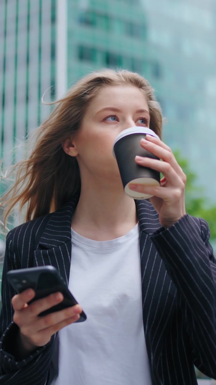 Ung forretningskvinde bruger smartphone og drikker kaffe udendørs.
