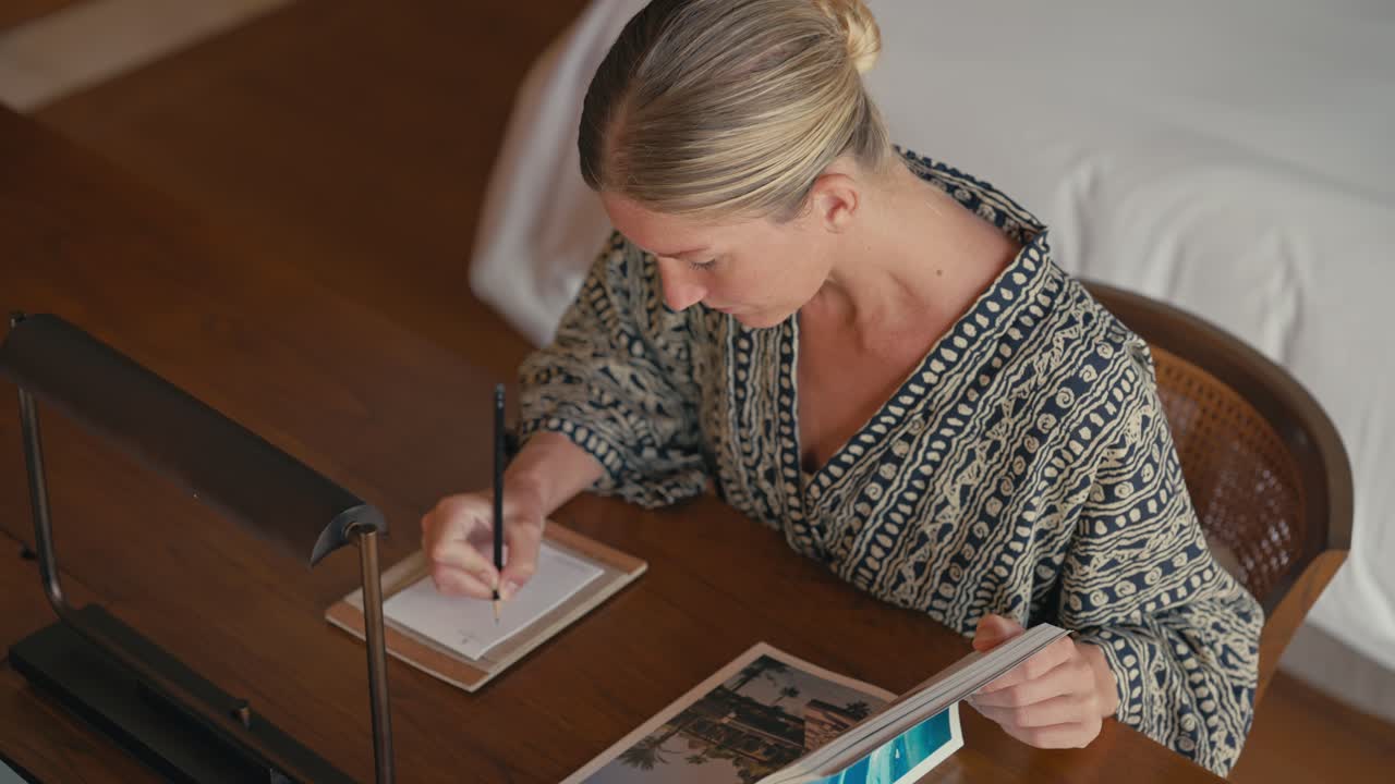 Donna caucasica bionda che prende appunti a una scrivania di legno mentre guarda una rivista in una villa resort di lusso.