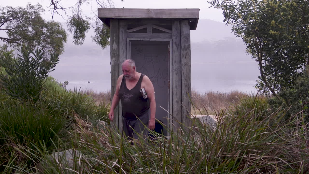 캐주얼 의상을 입은 남자가 기묘한 나무 화장실 밖에서 서서 팔 에 신문을 들고 있습니다.