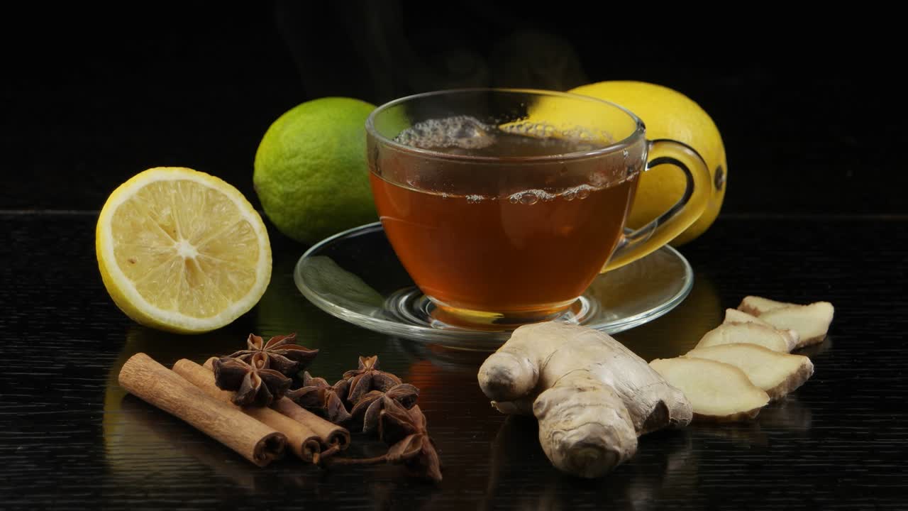 Steam rises above a cup of hot tea. Lemon, lime and spices for tea on a black wooden table