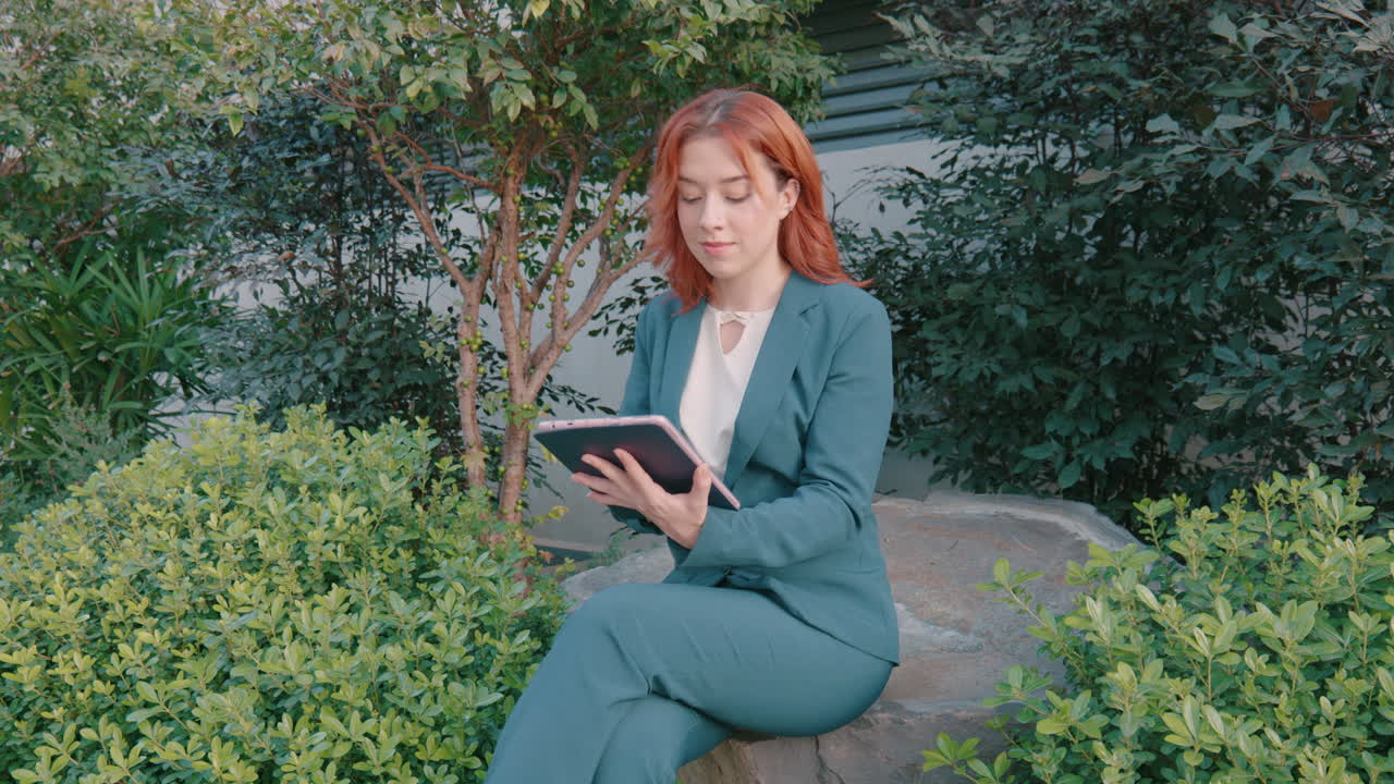 снимок красивой белой женщины, сидящей на скале в парке и использующей цифровой планшет для прокрутки приложений в ветреный день