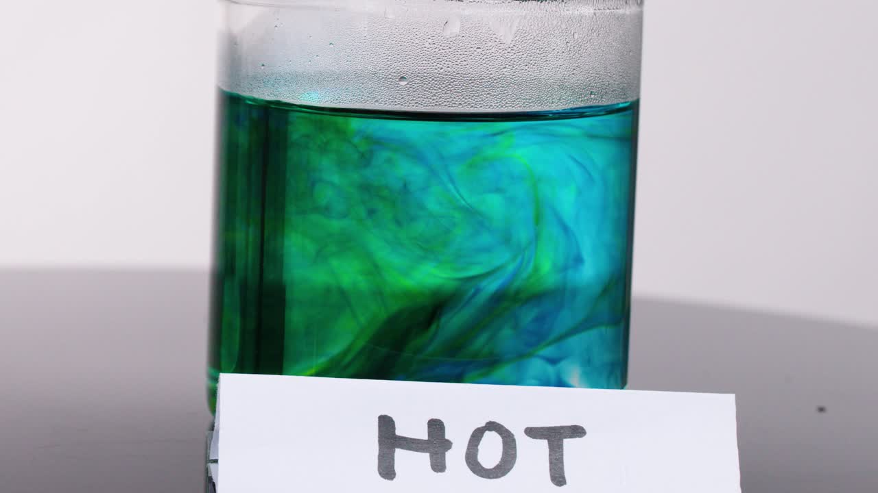 Green and blue food coloring disperses rapidly in hot water glass under bright studio lighting