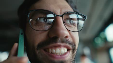 Close-up-of-a-happy-brunette-guy-with-stubble-in-glasses-communicates-on-a-green-smartphone-and-smiles-broadly-while-walking-around-the-office