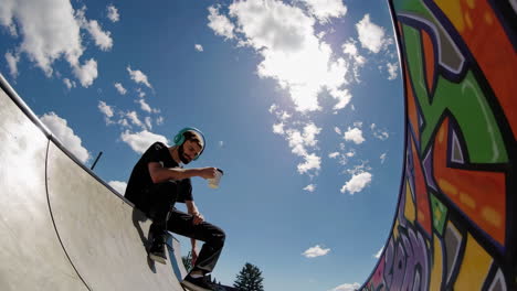 a man with headphones sits at a skate park under a sunny sky, holding a cup