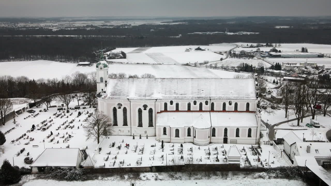4K drone pull-back reveals snow-covered Elchingen church amid fields and cemetery. Smooth ascent unveils winter beauty under clouds—ideal for promos, documentaries and tourism visuals
