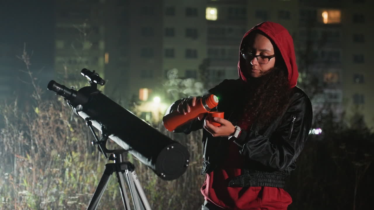 night rooftop stargazer adjusts telescope holding thermos, city lights glow behind apartment buildings, tripod set in tall grass, hooded observer calibrates eyepiece while breath fogs in cold air