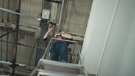 man in glasses and striped sweater standing on scaffold platform tightening lighting fixture on ceiling track system inside studio space, surrounded by cables and overhead mounting hardware during setup