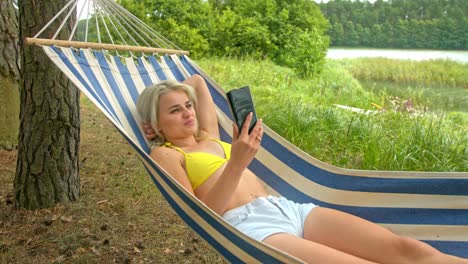 blonde girl lies on a hammock on a warm sunny summer day having a video call by cell phone