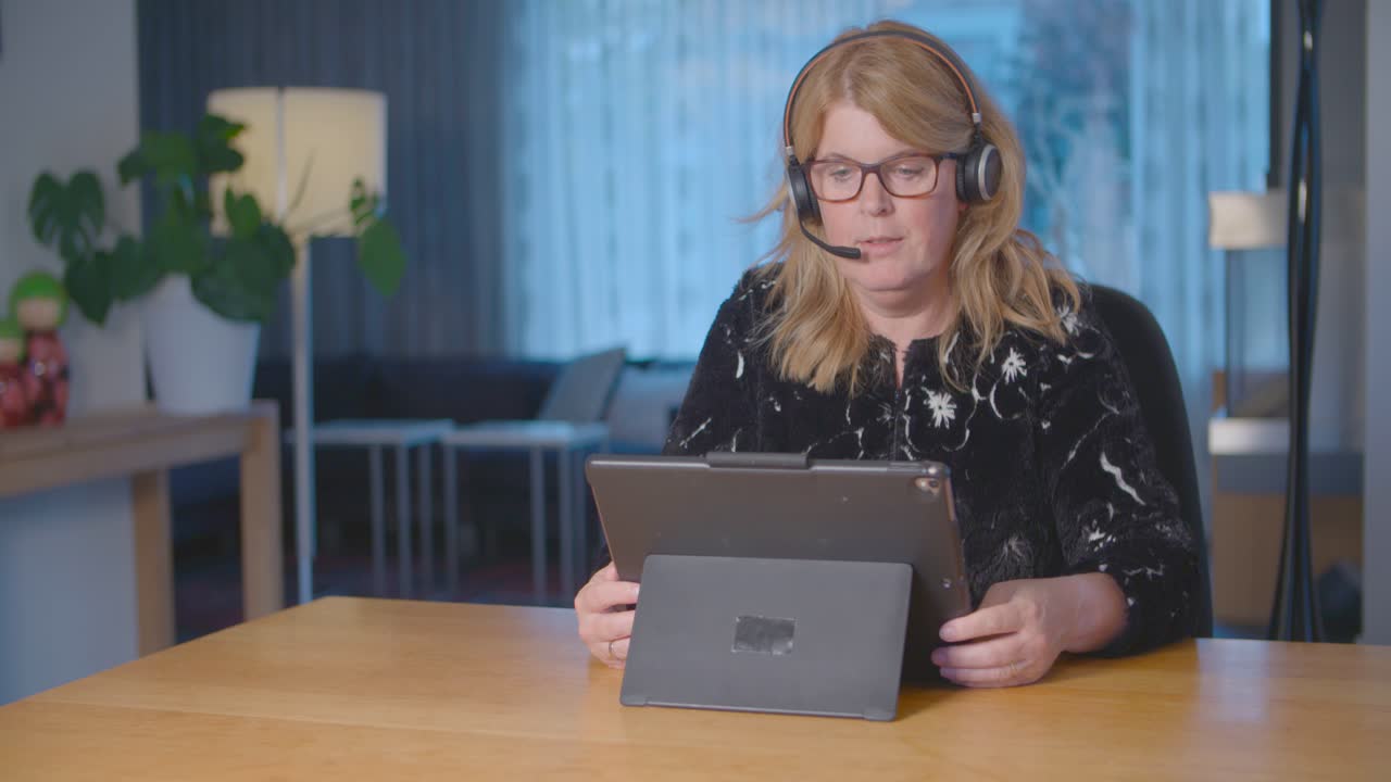 una mujer rubia llamando a sus colegas en una reunión de negocios con su tableta y auriculares mientras trabaja desde casa