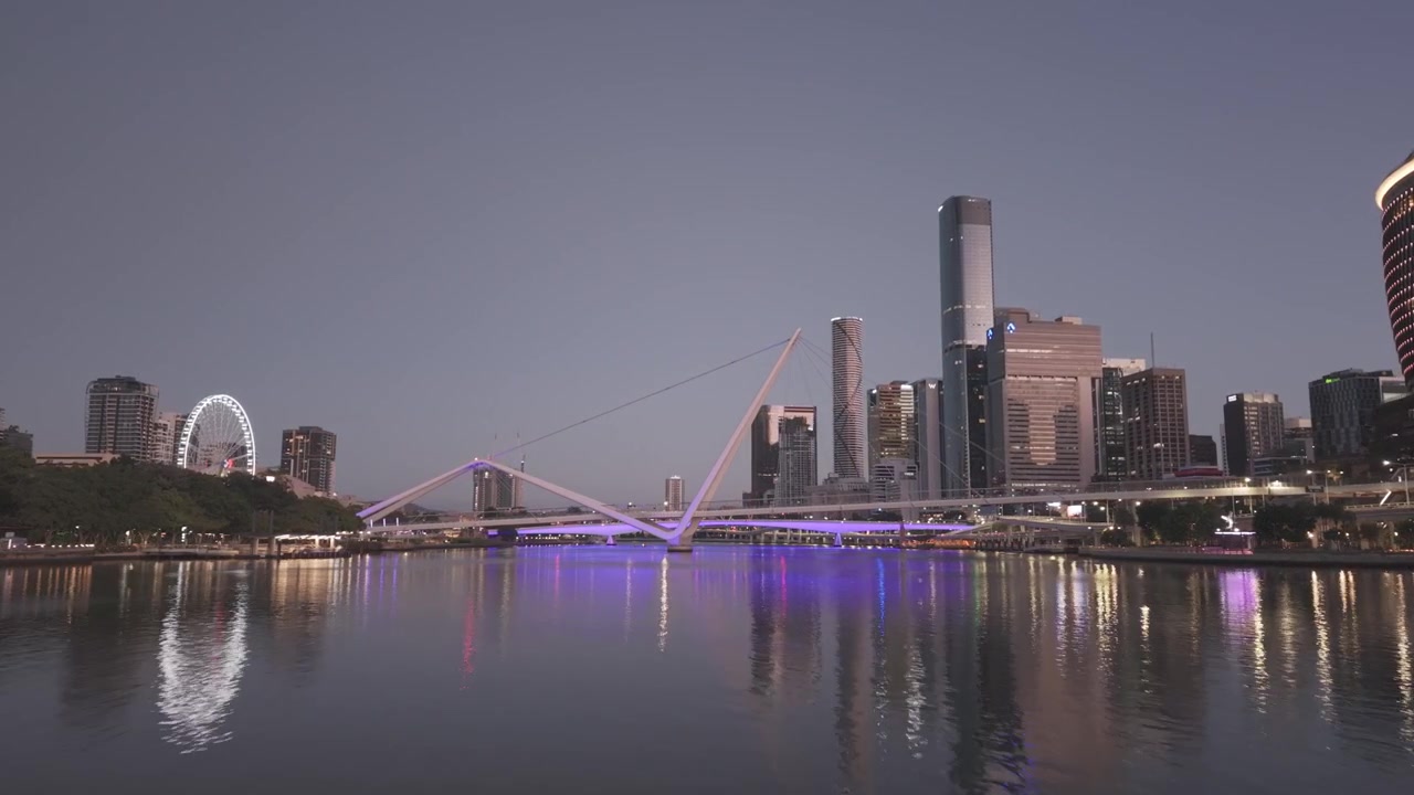 An aerial view at dusk shows the Brisbane riverfront skyline glowing above the calm, reflective river.