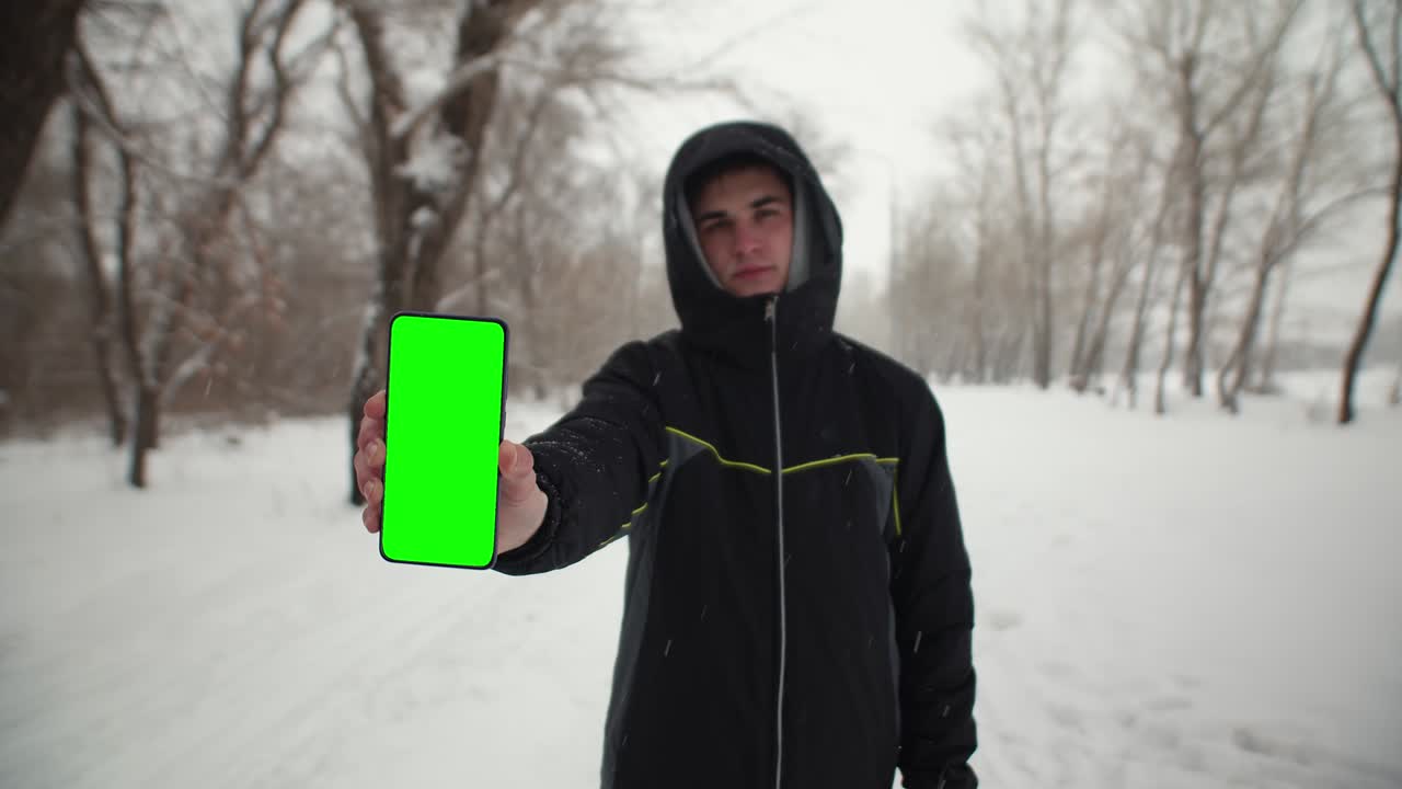 hombre en el parque invierno muestra pantalla de teléfono de chomakey. hombre sostiene un teléfono con una pantalla verde en la mano la naturaleza del invierno
