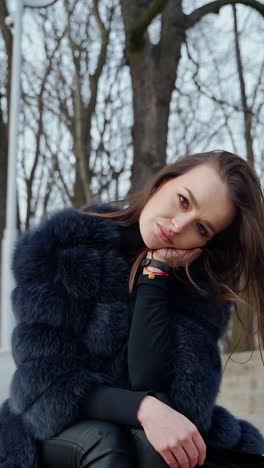 portrait of attractive model sitting in the park. beautiful young woman in black fur coat posing on camera outdoors. vertical video