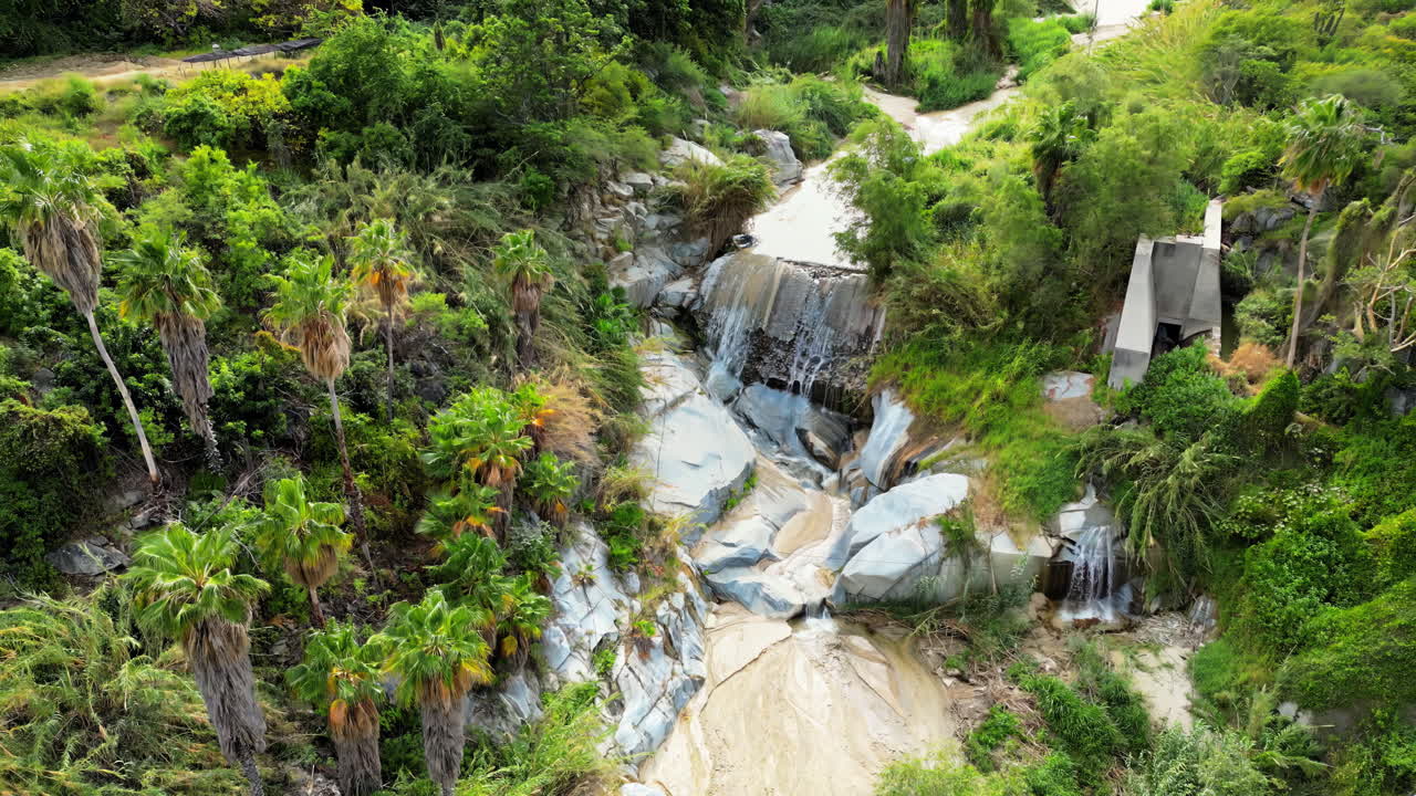 Aerial drone view of a small waterfall cascading over smooth rocks, surrounded by palm trees and wild plants in slow motion