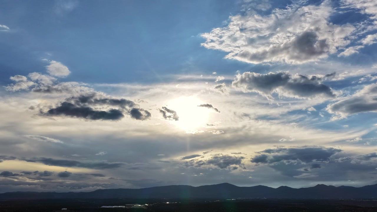 4K drone timelapse of the bright sun shifting behind scattered moving clouds. Dynamic sunbursts and lens flares streak across a vibrant blue sky above the Los Alamos, New Mexico landscape.