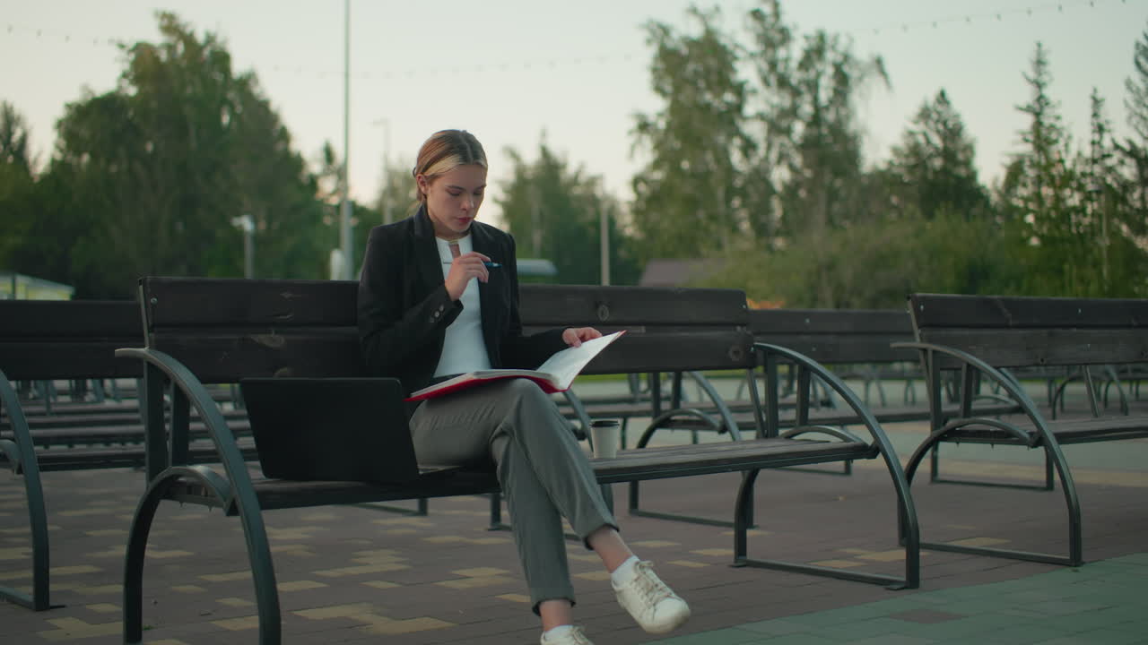 Woman seated alone on outdoor bench deeply focused on open file while holding pen with laptop and coffee cup by her side in quiet urban setting surrounded by trees with distant people and buildings