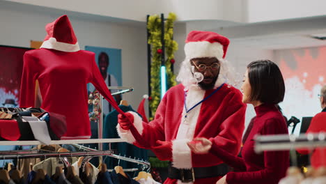 African-american-employee-dressed-as-Santa-Claus-helping-client-pick-festive-outfit-in-Christmas-shopping-store-during-winter-holiday-season.-Worker-assisting-customer-in-fashion-shop