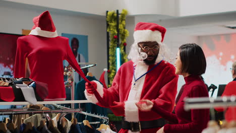 african american employee dressed as santa claus helping client pick festive outfit in christmas shopping store during winter holiday season. worker assisting customer in fashion shop