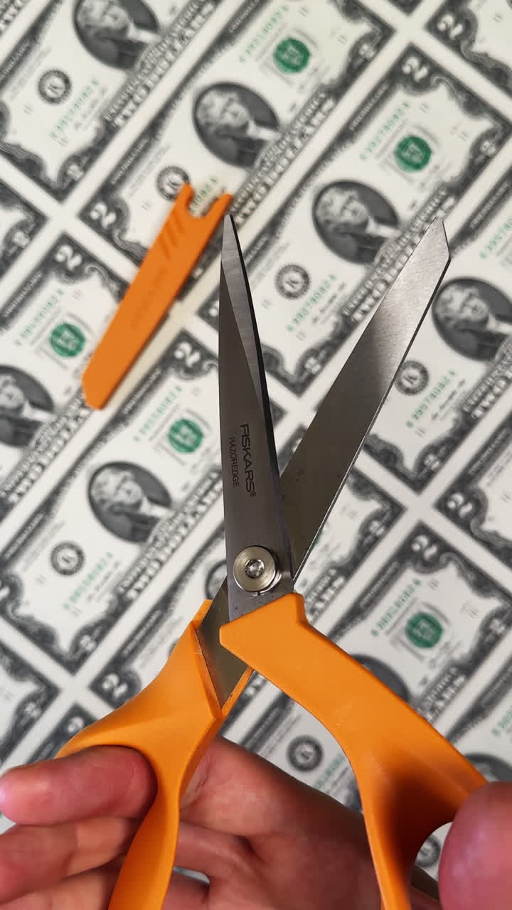 San Antonio, USA, 14 August 2025: Hand holds the scissors with orange finger loops. Close up. A large sheet of uncut two dollar bills at backdrop. Vertical video