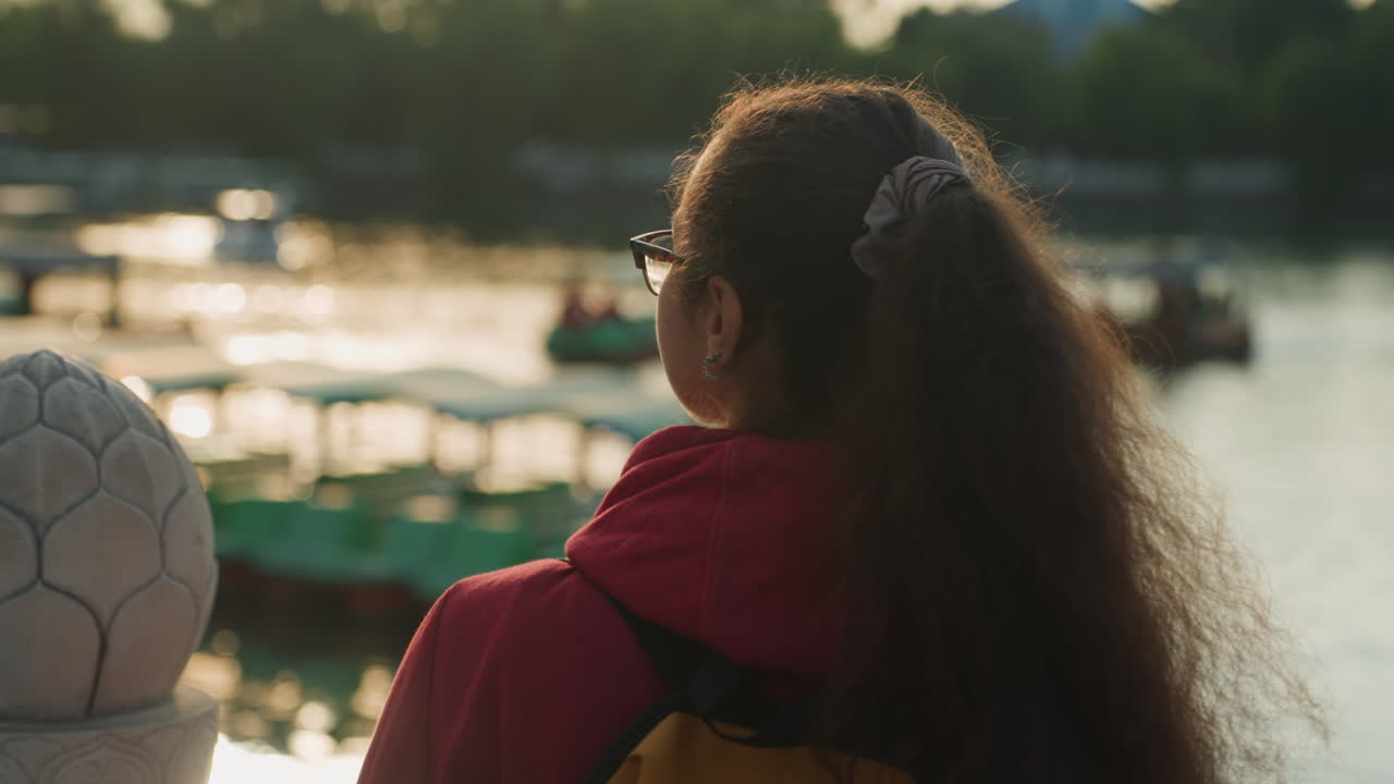 Young woman gazing at golden water, soft evening light highlights ponytail and glasses, backpack on shoulders, boats and marina shimmer, urban skyline blurred, contemplative mood, cinematic side