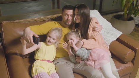 happy parents and two little girls taking selfie photo while sitting on sofa at home 1