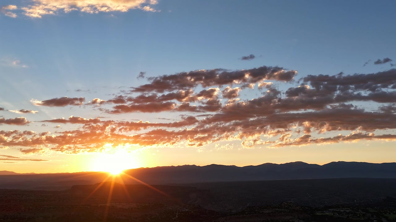 Timelapse de drone 4K UHD de um nascer do sol dourado no Novo México. O sol brilhante surge sobre a silhueta escura de uma montanha, projetando raios de sol nítidos para baixo através de uma camada de nuvens matinais dispersas e texturizadas