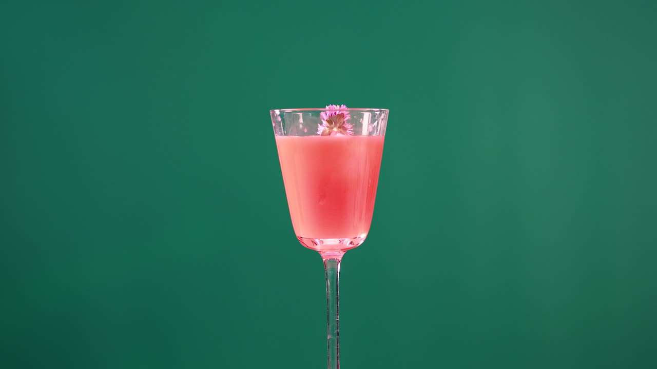 En lyserød cocktail pyntes med blomster ved hjælp af en tang mod en green screen-baggrund, hvilket fremhæver levende farver og præcise bevægelser