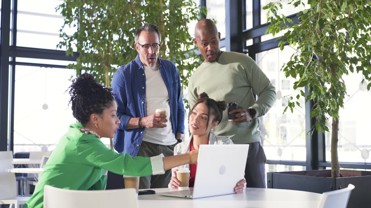 Diverse coworkers exchanging coffee leaning in pointing at laptop while working in office lounge