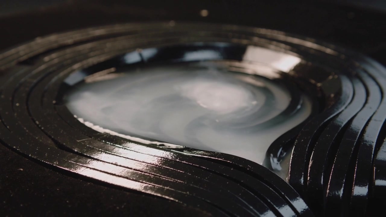 Spreading milky fluid forming spiral rings in center well of ridged black disc, studio filming