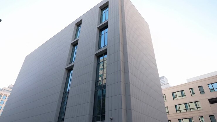 Uma vista de baixo ângulo que se move gradualmente para cima ao longo de um exterior cinza contemporâneo, enfatizando janelas estreitas com tonalidade azul, painéis de parede geométricos, uma linha de telhado nítida, edifícios próximos e um céu claro.