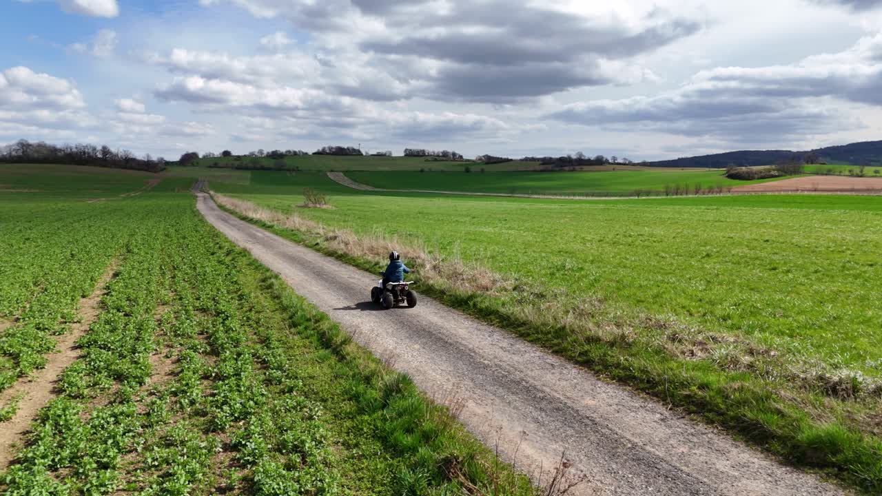 Ripresa statica con drone di un bambino che guida un quad lungo una strada sterrata rurale attraverso campi verdi, ampia vista aerea di un tranquillo paesaggio campestre.