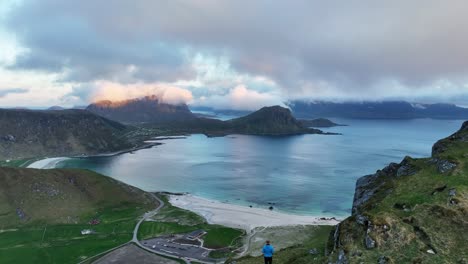 drone pulls back from haukland beach in midnight sun revealing man on cliff near mannen mountain
