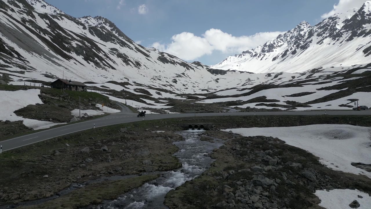 Aerial shot of snow-dappled Alps, a rushing stream, and a motorbike riding along a scenic curving road under a blue sky.