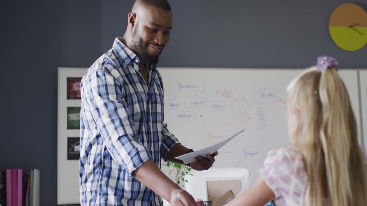 Video of happy african american male teacher giving test an clapping hands with caucasian girl