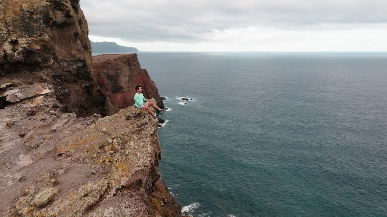 Aerial orbit shot of a man seated on a cliff edge, gazing at the ocean and rugged cliffs of Ponta de São Lourenço.