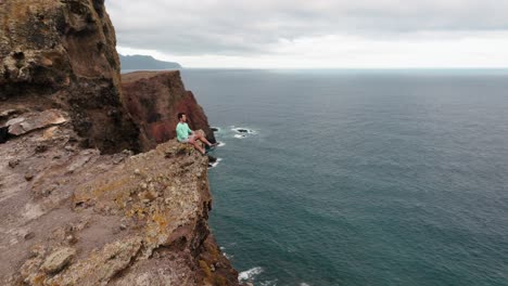 aerial orbit shot of a man seated on a cliff edge, gazing at the ocean and rugged cliffs of ponta de são lourenço.