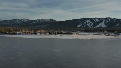 Aerial-flyover-frozen-lake-towards-remote-village-with-mountain-landscape-in-the-background