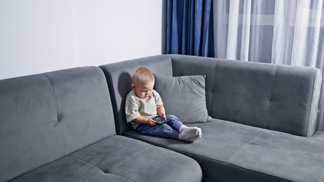 Sød toårig baby ser opmærksomt på smartphonen. Lille barn spiller et spil på telefonen. Småbarn ser tegnefilm siddende på sofaen med telefonen i hænderne