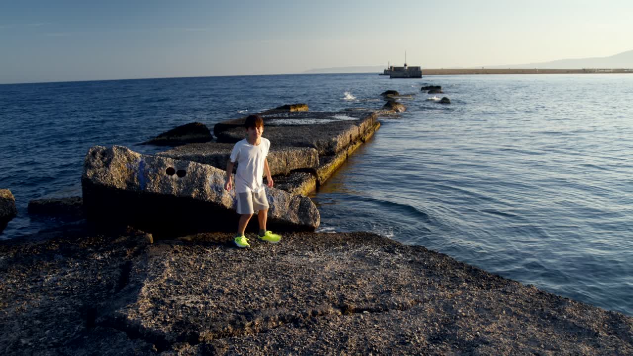 Medium shot of 8 years old caucasian boy, walking towards the sun on a pier at sea Kalamata, enjoying his summer vacations 4K
