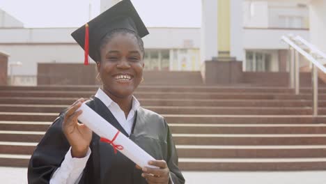 young african american female graduate standing in front of the camera with a diploma and books in her hands. the student is dressed in a black robe and a square master's hat and stands outside.