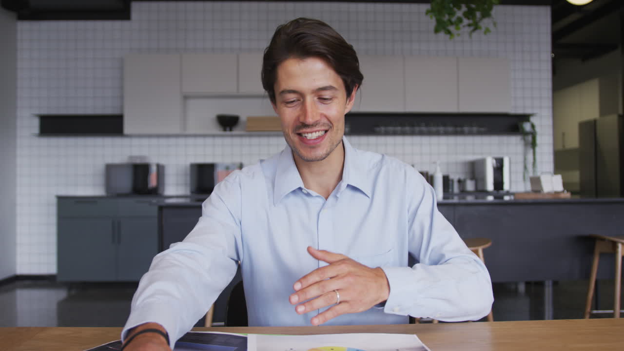 Uomo d'affari caucasico sorridente in videochiamata che esamina documenti nella cucina dell'ufficio