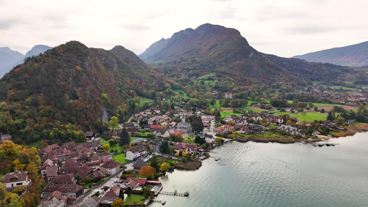 Aerial perspective of Chateau de Duingt, France, featuring a lakeside village bordered by rolling mountains, winding roads and autumn forests with varied foliage tones and calm water