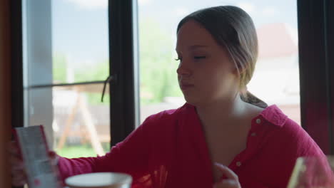 young brunette woman in crimson shirt sits at wooden table near window, leaning forward searching tabletop for lost item while yard outside glows, ruby drinks and glass pitcher blur in foreground