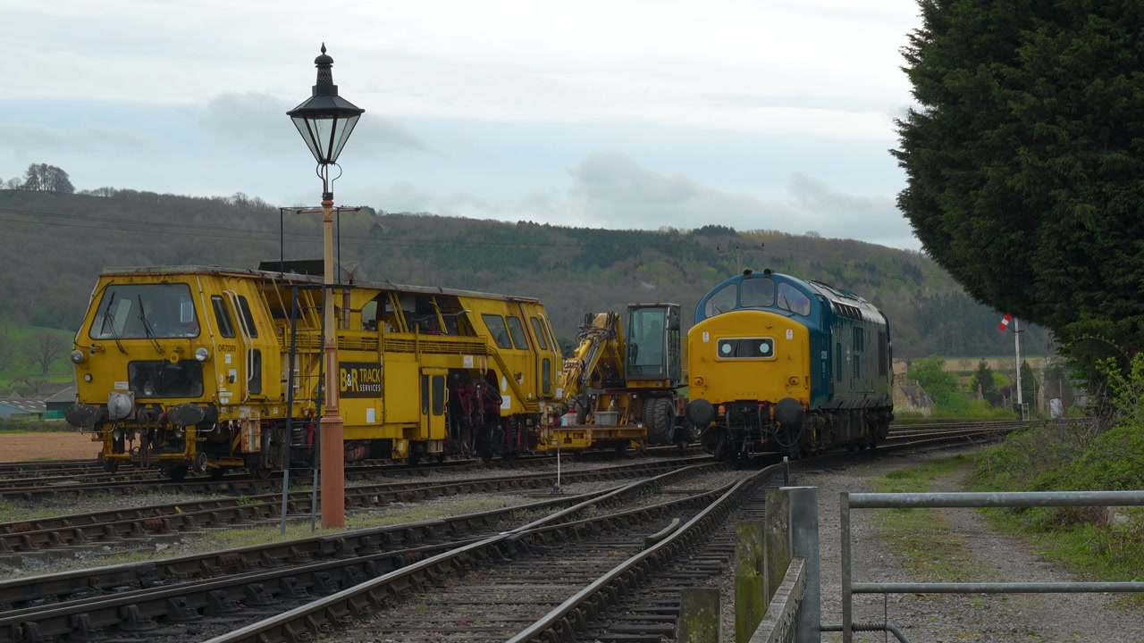 Una classica locomotiva diesel blu e gialla supera un veicolo giallo per la manutenzione dei binari su binari paralleli in un deposito ferroviario rurale inglese.