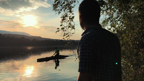 Backlit scene as a man on shore watches a canoe pass by against a golden sunset