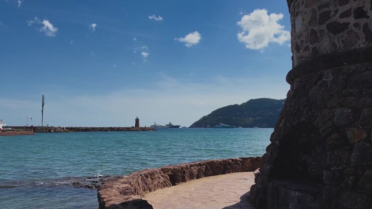Un bastione storico in pietra si affaccia sull'acqua turchese del Mediterraneo, su un ingresso del porto, su barche lontane e su verdi colline costiere sotto un cielo azzurro brillante.