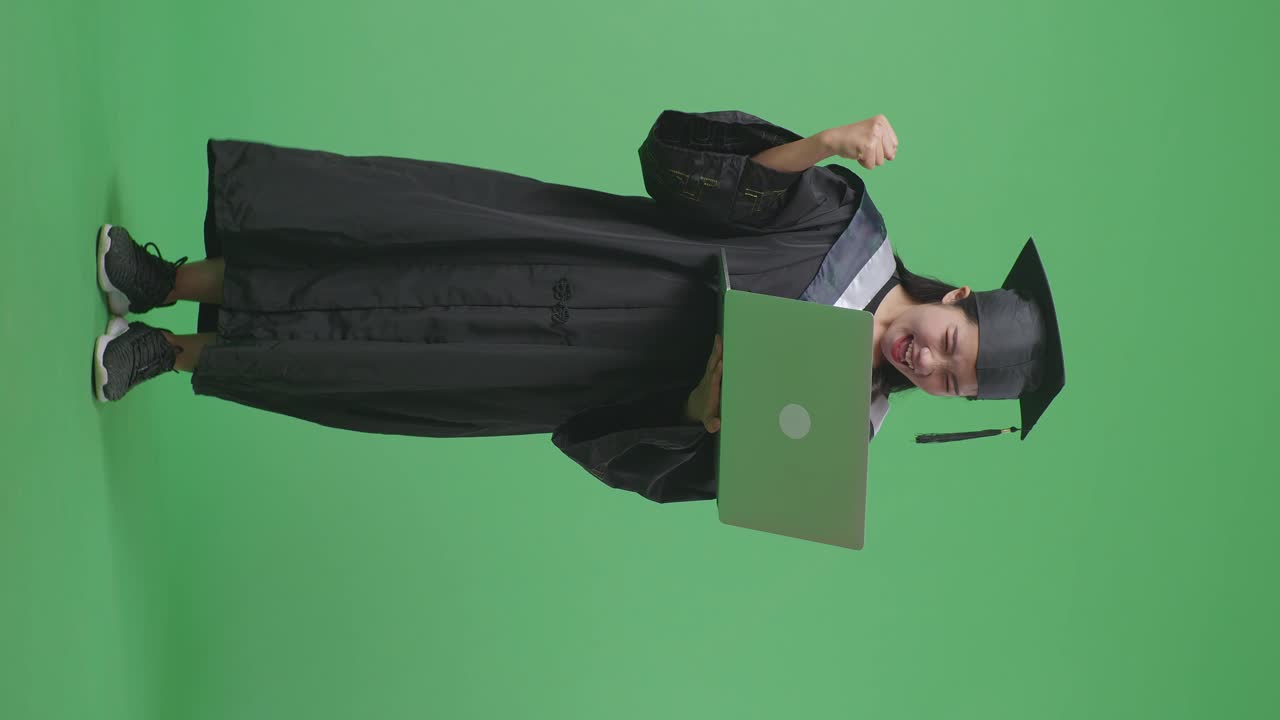 Studentessa asiatica laureata a figura intera, con tocco e toga, che guarda un portatile in mano e poi esulta, su sfondo a schermo verde in studio