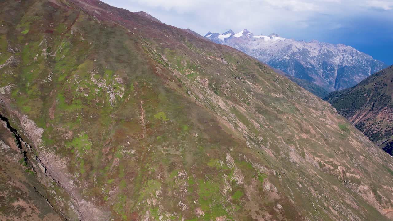 Aerial shot of steep green-brown mountain slopes with snowy peaks in the distance. Kashmir